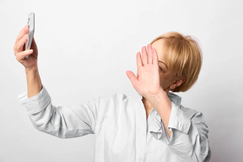 Woman smartphone selfie hand face privacy studio white portrait gesture, playful closeup, a person covers eyes while holding a smartphone, blank white studio background, soft light