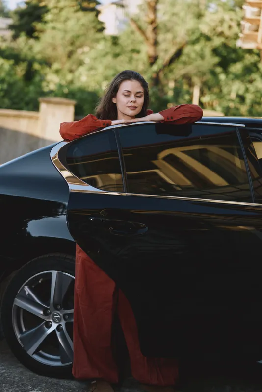 Young woman in a red dress rests against a sleek black car outdoors, eyes closed, a peaceful moment captured in soft light, with a subtle breeze and warm tones creating a relaxed lifestyle vibe
