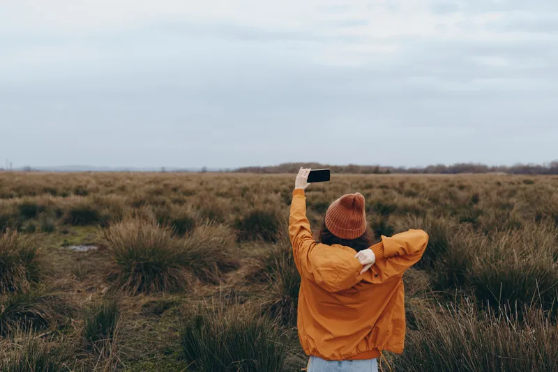 Woman outdoors in an orange jacket and beanie, standing on a grassy field path, lifting a phone to take a photo, landscape horizon view, calm lifestyle, cloudy sky, rural travel adventure