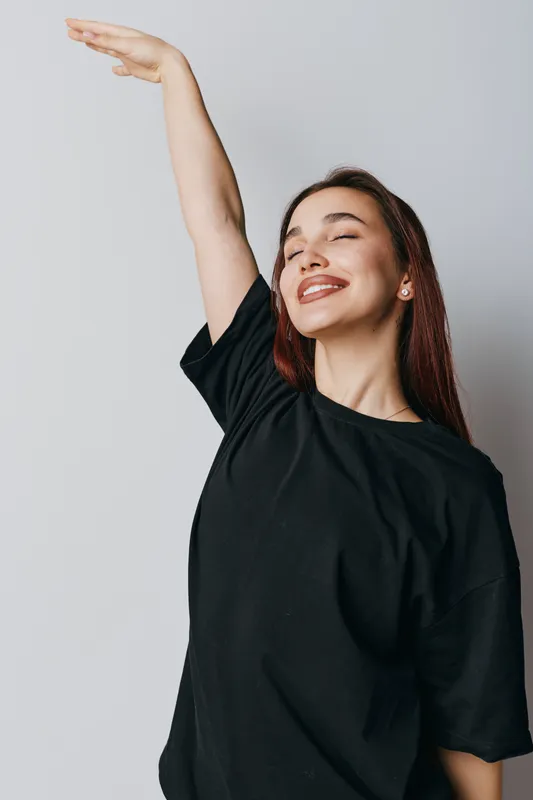 Young woman with long hair and closed eyes smiling happily while raising one arm up wearing a black t shirt on plain light background. Positive emotion and freedom concept.