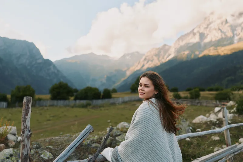 Young woman with long curly hair wrapped in a blanket stands near a wooden fence in a mountain valley during a calm day with soft sunlight.