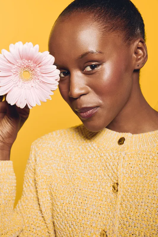Close-up of a smiling African woman with dark skin holding a pink flower near her face. She wears a yellow knitted sweater against a bright yellow background. Concept of beauty and nature.