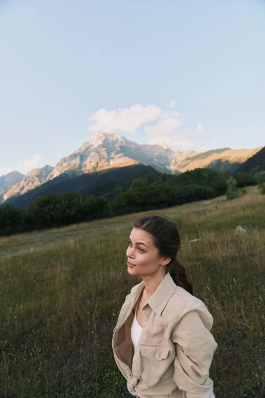 A woman in a beige jacket stands in a grassy field with distant mountains, looking to the side. Natural mood for outdoor lifestyle and calm moments.