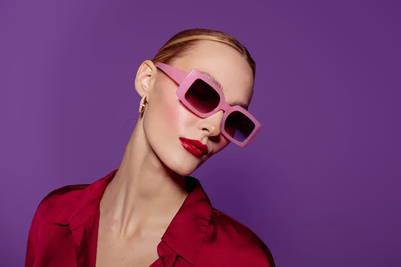 Stylish woman with slicked hair wears pink sunglasses and red lipstick. She poses against a purple background wearing a red shirt. Concept of fashion, beauty, and modern style.