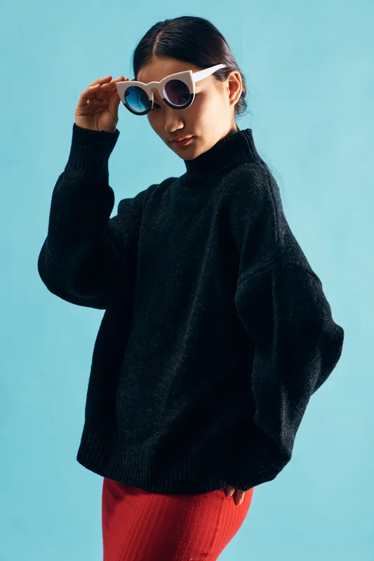 Woman wearing white sunglasses and black turtleneck sweater stands against blue background, posing with hand near face and wearing red pants, fashionable and stylish portrait.