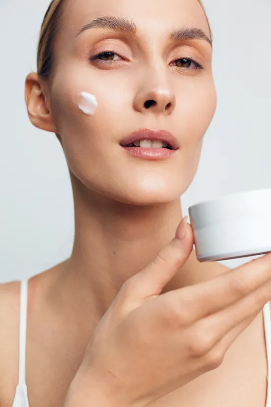 Woman skincare cream face studio beauty applying jar minimal white background, young woman with fresh expression, applying moisturizer on cheek, smooth complexion, clean aesthetic