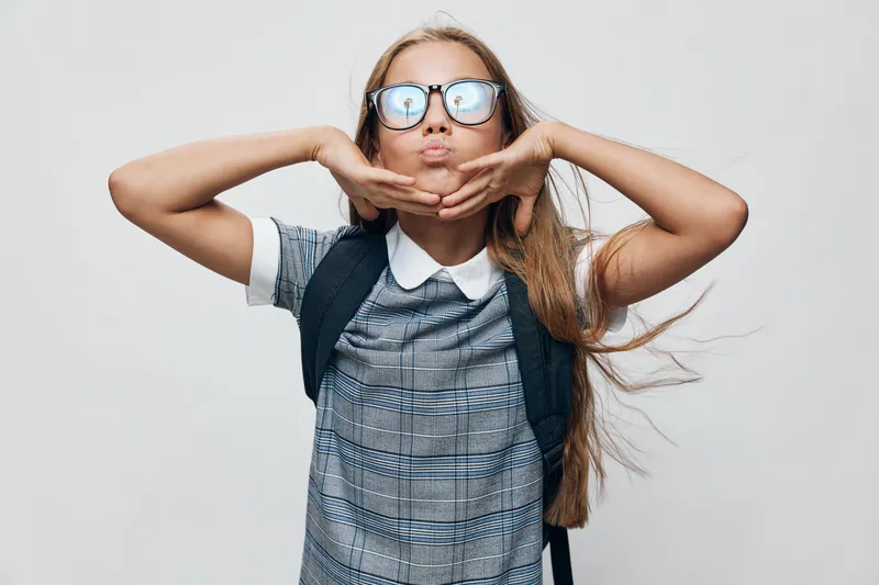 Girl with glasses and backpack making funny face and hands under chin posing on gray background. Young female student, smiling and playful in casual clothing with long hair.