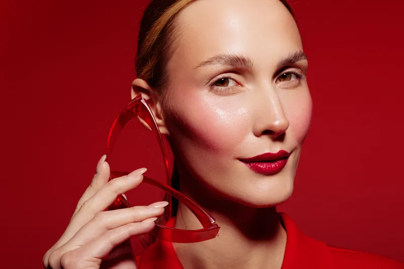 Close-up of a confident woman with glossy skin and red lipstick holding red sunglasses near her face. Red background and clothing create a bold and stylish fashion portrait concept.