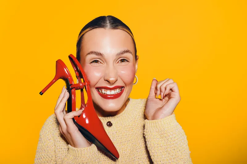 Smiling woman with red lipstick holds red high heel shoe near face on bright yellow background. Concept of fashion, style, happiness, and beauty with vibrant colors and joyful mood.
