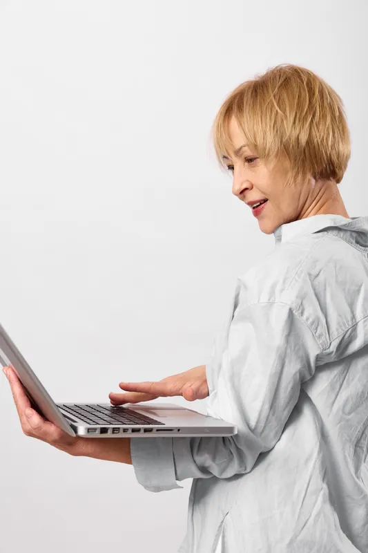 Woman elderly laptop typing working technology communication portrait indoors white background, senior female holds laptop, hands on keyboard, smiling while working, remote internet communication on