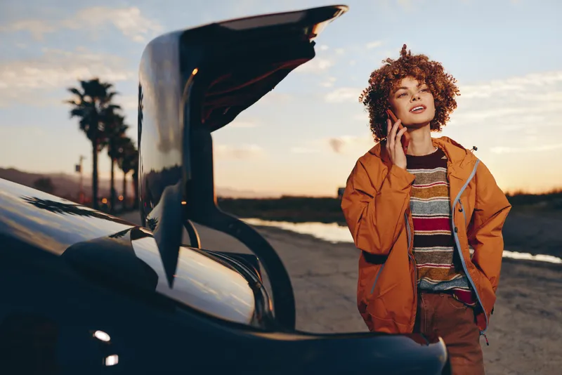 Woman lifestyle scene with curly hair, orange jacket, beside an open car trunk at sunset, backpack in hand on an outdoor road. Travel moment with warm soft light for adventure.