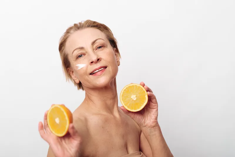 Mature woman with short hair holds two halves of fresh orange, smiling with natural skincare cream on cheek. Concept of healthy nutrition, beauty, wellness, and youthful skin care.