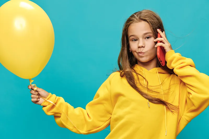 Girl in yellow hoodie holding balloon and talking on mobile phone isolated on blue background for communication, childhood, fun, technology and casual lifestyle.