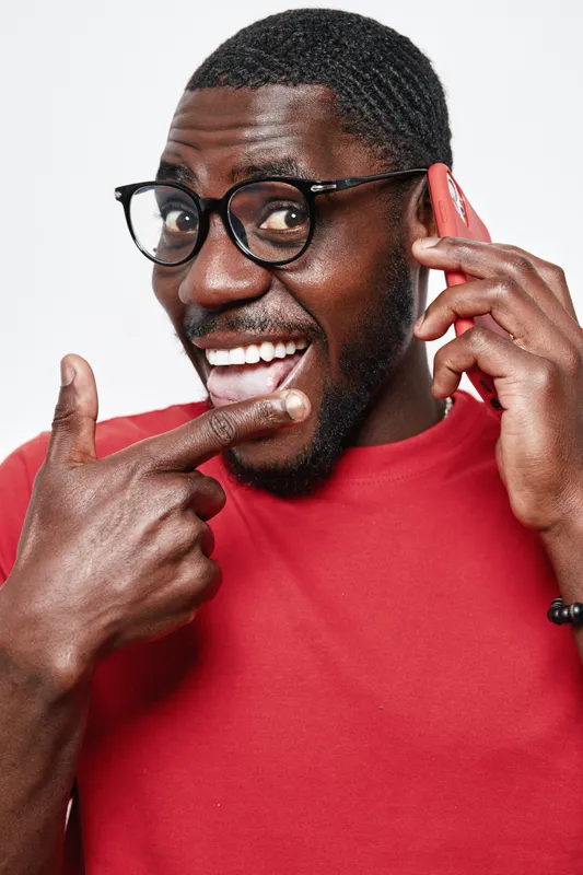 African American man with dark skin wearing glasses and red shirt talking on smartphone smiling and pointing at his face with excitement and happiness on white background.