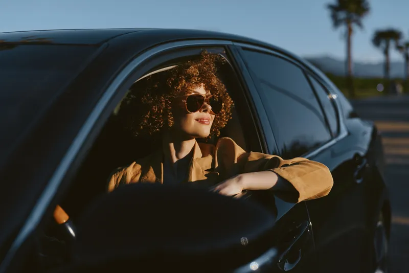 Woman driving a car in warm retro film color, wearing sunglasses and brown jacket, relaxed urban mood, sunlit interior, candid travel moment, stylish road trip vibe