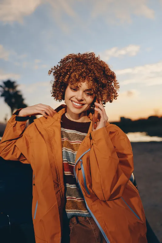 Woman lifestyle outdoors, golden sunset, soft breeze, smiling, curly hair, orange jacket, hands near ears, warm evening light, relaxed posture, scenic sky, gentle distant palms