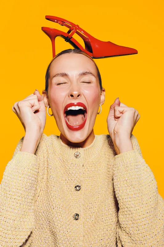 Excited woman with red lipstick and earrings wears yellow sweater and balances red high heel shoe on head. Bright orange background creates energetic and playful mood in studio portrait.