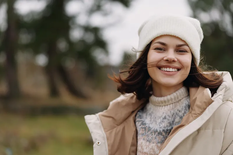 Woman in an outdoor setting wearing a beige jacket and knit beanie, smiling into the camera with a soft autumn breeze, natural light, casual fashion portrait in a park, candid and warm.