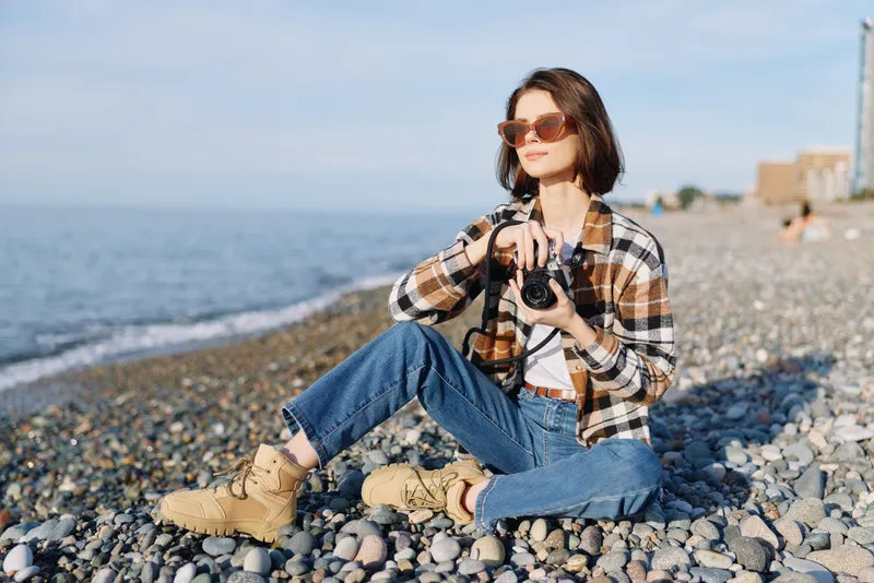 A woman sits on a pebbled beach in jeans and boots, holding a camera with a relaxed pose, enjoying seaside light and scenery, calm mood and outdoor photography vibe.