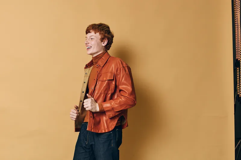 Young man wearing orange leather jacket and dark pants standing and smiling against beige background with casual style and red hair in studio environment.