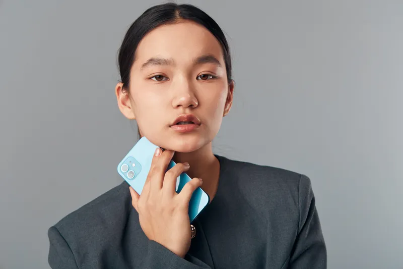 Young woman holding smartphone near face looking at camera with neutral expression wearing gray blazer on gray background in studio for technology and communication concept.