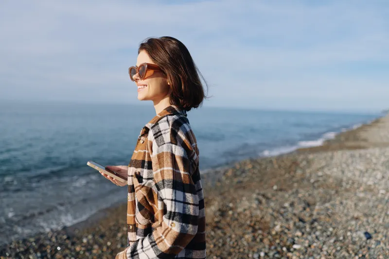 Woman by the sea wearing sunglasses and a plaid shirt, holding a smartphone, enjoying a breezy coastal moment in daytime light.
