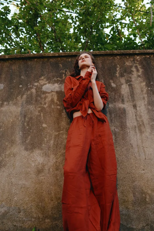 A woman in a red jumpsuit leans against a weathered wall in an outdoor setting, expressing confidence, style, and street fashion in a candid, contemporary moment