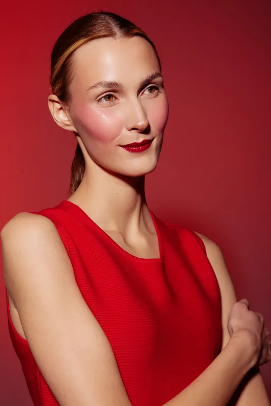 Portrait of a confident woman in a red sleeveless top posing against a red background. She has natural makeup with glossy lips and a subtle smile, expressing elegance and style.