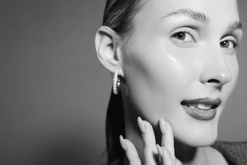 Close up of a young Caucasian woman with fair skin touching her face, looking confident with earrings on a grey background. Concept of skincare and natural beauty.
