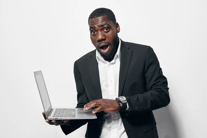 Black man with dark skin in business suit holding laptop with surprised expression on white background. African ethnicity, professional, technology and communication concept.