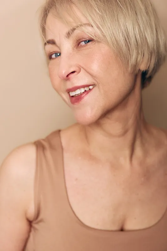 Close up of smiling mature woman with short blonde hair wearing beige tank top against neutral background. Concept of natural beauty, aging gracefully, and confident female portrait.