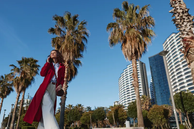 Woman outdoors in a red coat, smiling and confident, sunny day among palm trees and city skyscrapers, fashion forward urban moment with vibrant colors