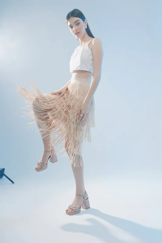 Young woman in a fashionable beige fringe skirt and white crop top poses against a light background. Concept of woman beauty fashion and modern style.
