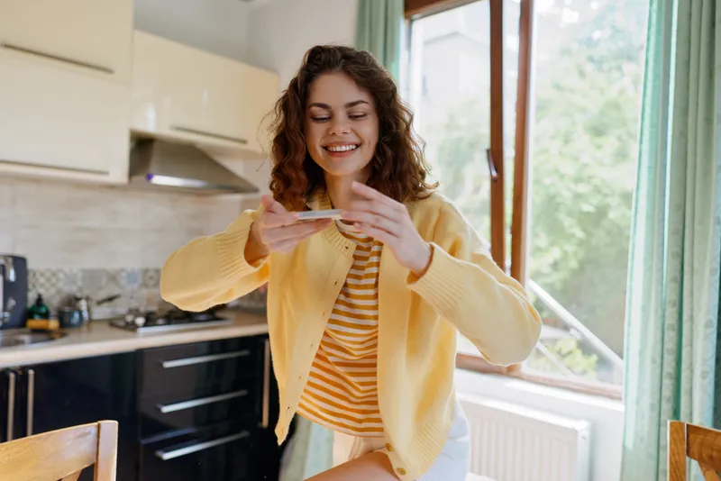 Cheerful young woman in a yellow cardigan smiles at her phone in a bright home kitchen during a casual moment.