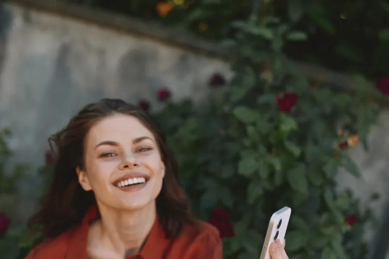 Smiling woman with long hair outdoors, holding a smartphone. Casual lifestyle scene showing happiness, connection, and everyday moments in natural daylight and warmth for a welcoming mood