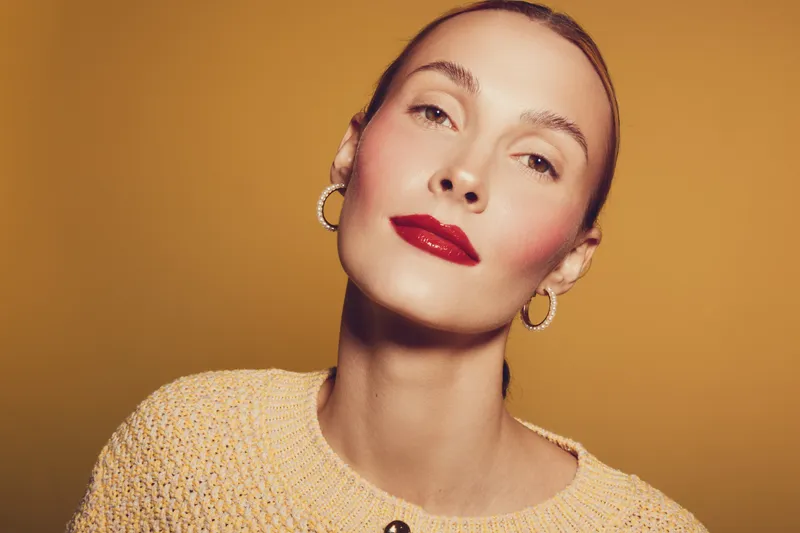 Portrait of confident woman with red lipstick and hoop earrings wearing knitted sweater on yellow background. Concept of beauty, fashion, and elegance with warm studio lighting.
