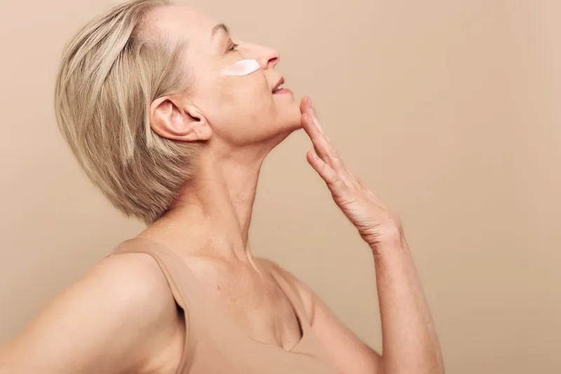 Mature woman with short blonde hair applies face cream on cheek, eyes closed, enjoying skincare routine. Beige background and soft lighting create calm self-care concept.
