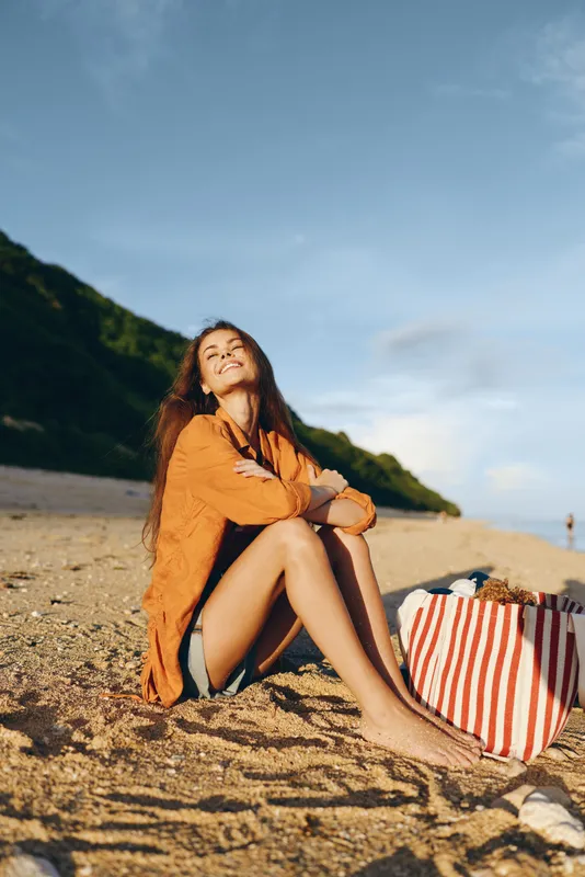 Young woman with long brown hair enjoys a sunny day sitting barefoot on a sandy beach near green hills with a striped bag beside her.