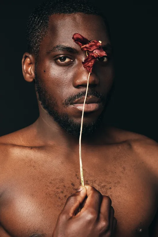 Creative conceptual portrait of African man holding dried flower in front of face with serious expression, dark background, artistic visual style, closeup and emotional mood.