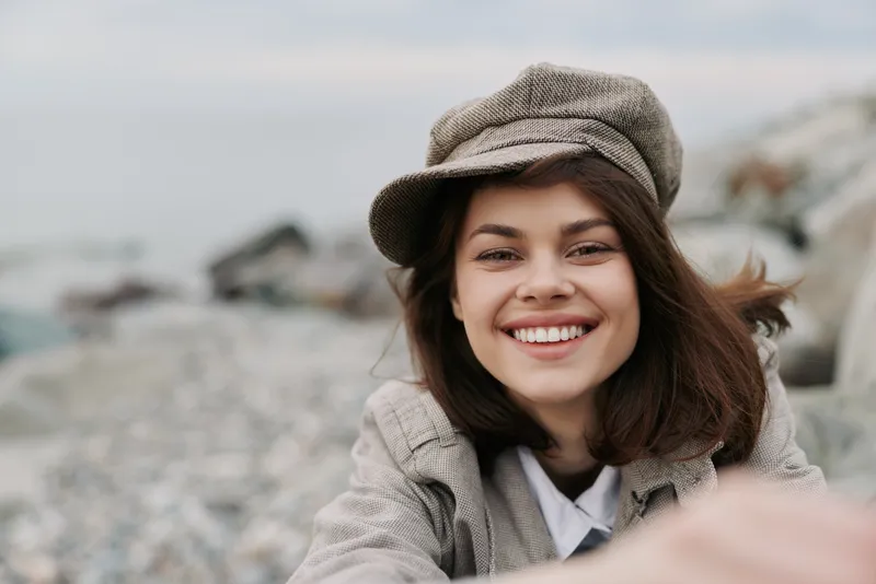Smiling young woman with brown hair wearing a beige cap and coat outdoors near rocks. Natural light highlights her joyful expression. Concept of happiness and casual fashion.