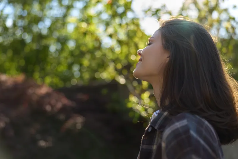A woman in a plaid shirt stands outdoors, gazing upward with a calm, thoughtful expression among sunlit trees, embodying outdoor lifestyle and mindfulness