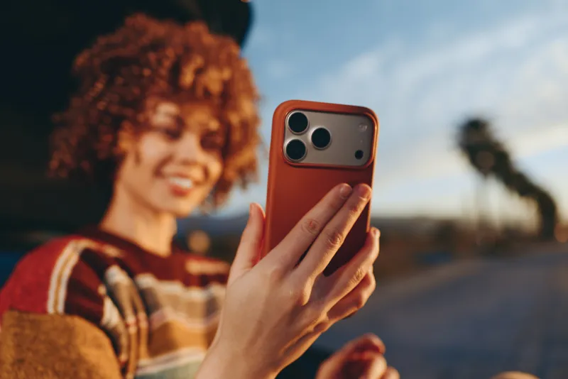 Woman lifestyle selfie outdoors, smiling with curly hair, holding smartphone up in hand, warm soft sunset light, relaxed travel moment, shallow depth of field, blurred road background