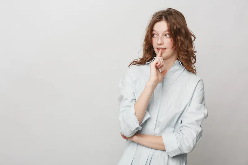 Thoughtful young woman with curly hair wearing a white shirt looks sideways with finger on lips. Concept of contemplation, curiosity, and quiet reflection in casual studio setting.