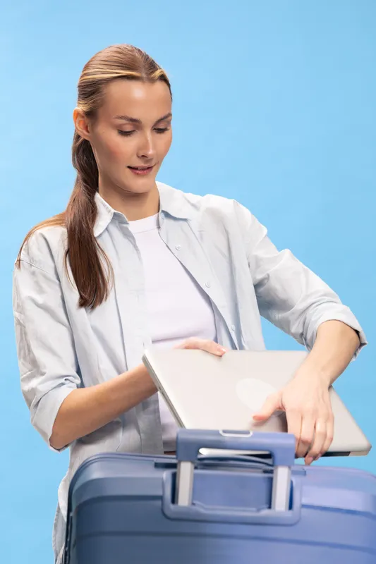Young woman packing a laptop into blue suitcase on blue background. Concept of travel preparation, business trip, and casual lifestyle with modern technology and light clothing.