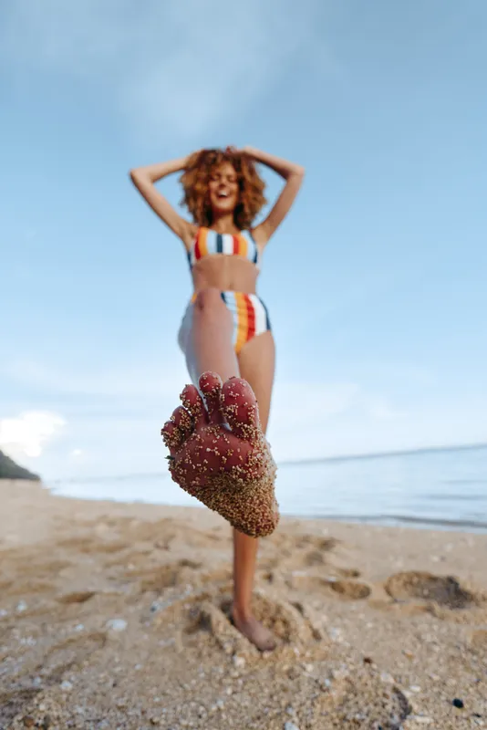 Woman beach bikini pose, holding a heart made of sand, smiling toward camera on a sunny shore, ocean waves and blue sky behind, summer vacation mood, natural light, soft breeze, relaxed stance