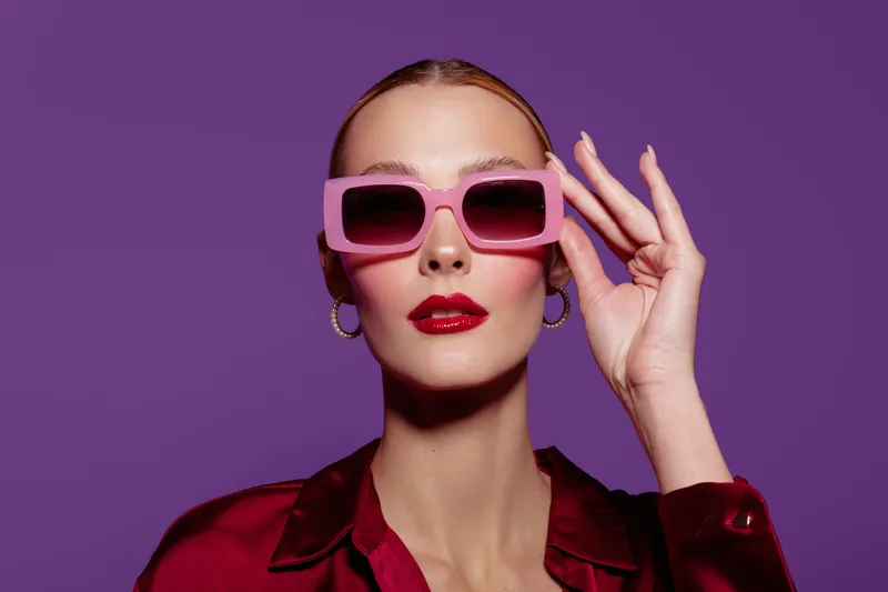 Stylish woman with pink sunglasses and red lipstick poses confidently against purple background. Fashion concept with bold colors and modern accessories for trendy look.