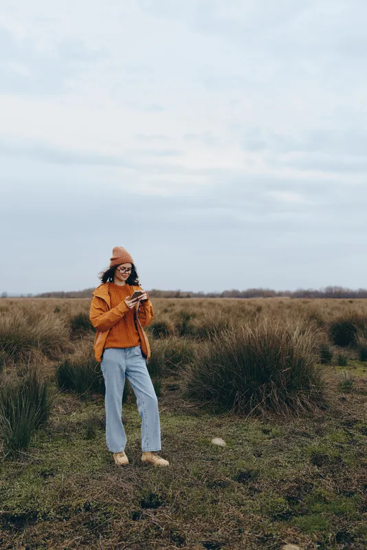 Woman lifestyle outdoors in nature, standing in a field of grass on a cloudy day, wearing an orange jacket and warm scarf, calm pose, open sky background.