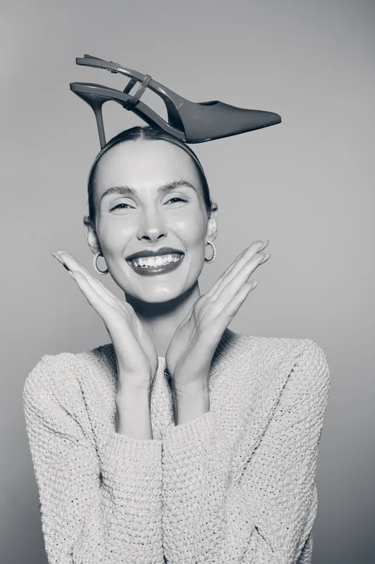 Smiling Caucasian white woman with dark hair wears high heel shoe on head, happy face and joyful mood.