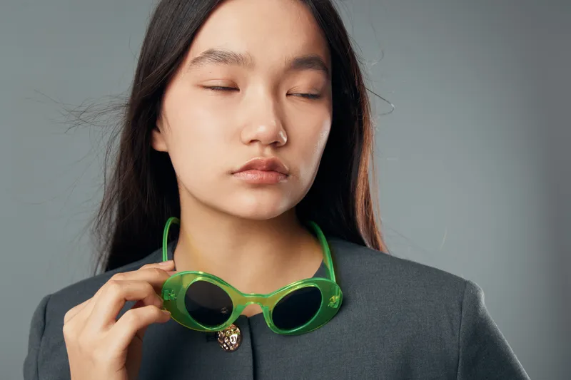 Young woman with long hair holding green sunglasses close to face with eyes closed in studio. Casual fashion, portrait and calm expression on grey background for lifestyle and style.