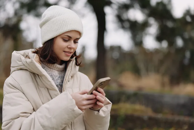 Winter scene shows a woman in a warm coat using a smartphone outdoors, with a calm natural backdrop, highlighting casual lifestyle, technology and communication on a chilly day.